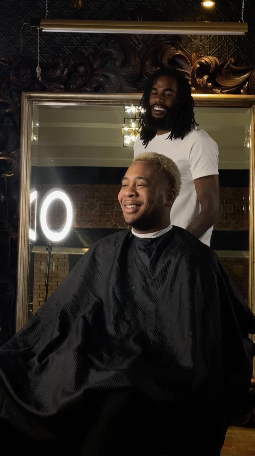 Jayy Klipsz laughing with a client in the chair in front of the gold framed mirror after a finished blonde taper